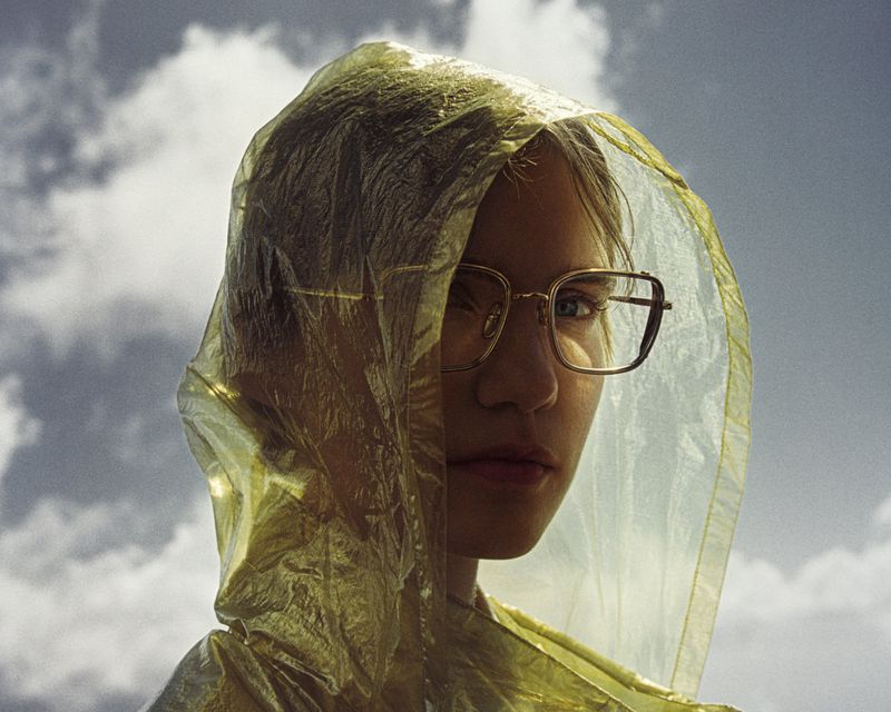Close-up of a young adult wearing a clear yellow plastic raincoat with hood up and large glasses, set against a cloudy sky.