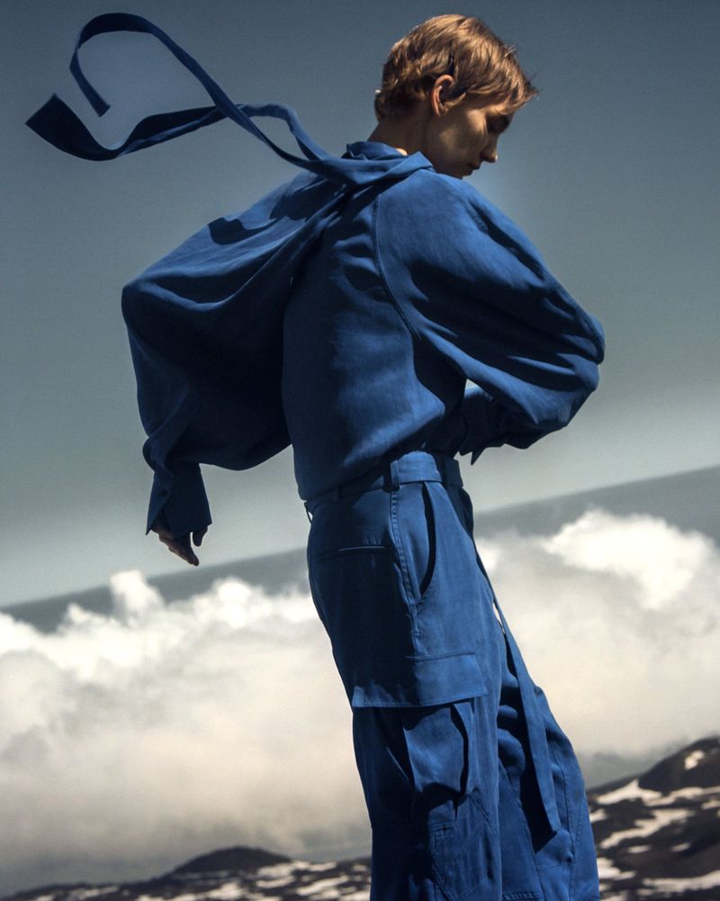 Fashion shot of a person wearing a blue outfit with an oversized jacket and billowing sleeves, posing outdoors against a mountain background.
