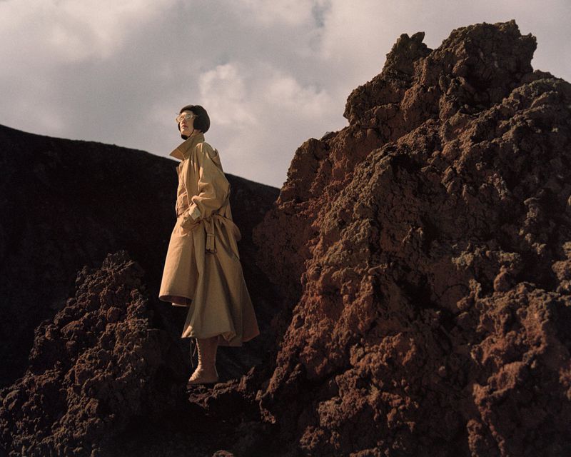 A person in a tan coat stands on rocky terrain under a cloudy sky. The focus is on fashion and the natural environment.