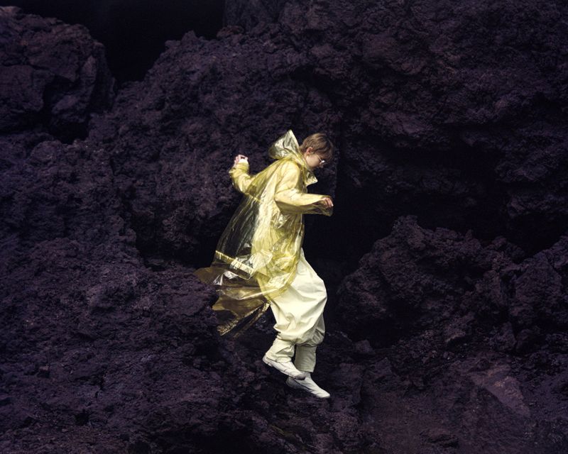 Young adult in yellow raincoat leaps down rocky terrain in outdoor fashion editorial, showcasing texture, depth, and muted color palette.