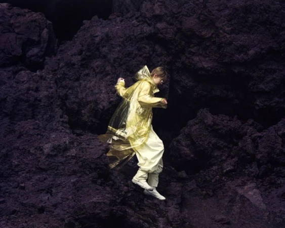 Young adult in yellow raincoat leaps down rocky terrain in outdoor fashion editorial, showcasing texture, depth, and muted color palette.
