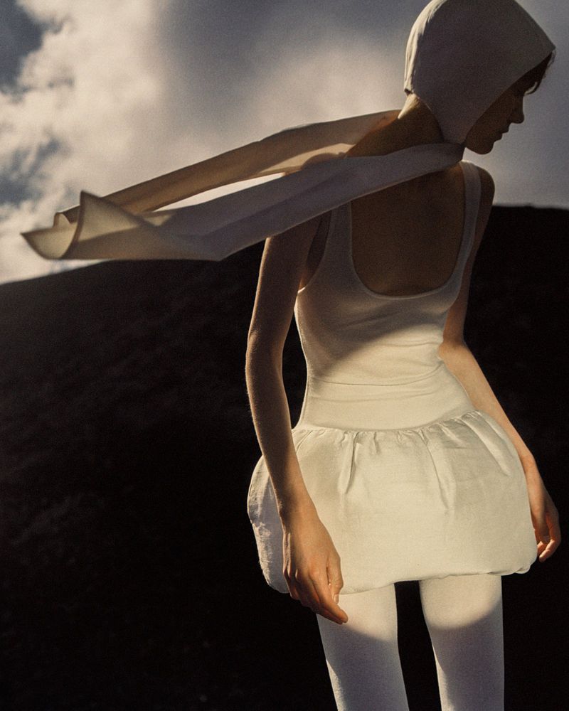 Woman in white hooded top, sleeveless shirt, and layered skirt poses outdoors against a dark hilly landscape beneath a cloudy sky.