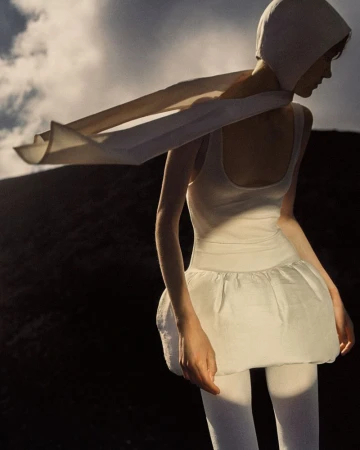 Woman in white hooded top, sleeveless shirt, and layered skirt poses outdoors against a dark hilly landscape beneath a cloudy sky.