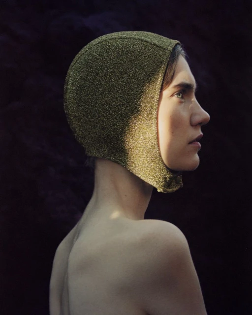 Adult female model poses with a golden hood, showcasing a side profile in a studio setting against a dark, textured backdrop.