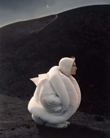 Fashion shot of a person in an oversized white puffy jacket on a black rocky surface near a mountain with a lava flow.