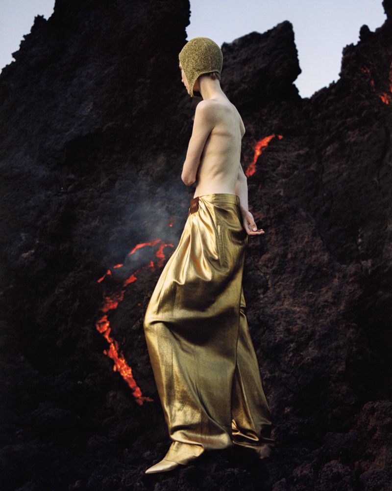 An adult wearing a gold-colored cap and long skirt walks on black lava rocks. There is smoke or steam rising from the rocks.