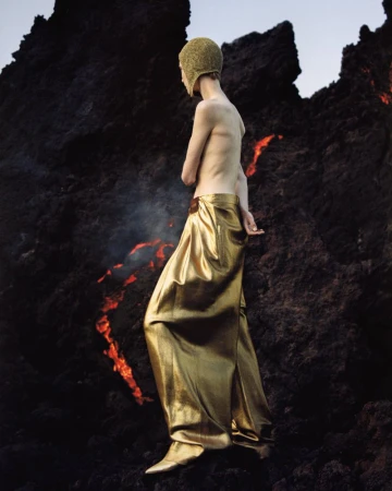 An adult wearing a gold-colored cap and long skirt walks on black lava rocks. There is smoke or steam rising from the rocks.