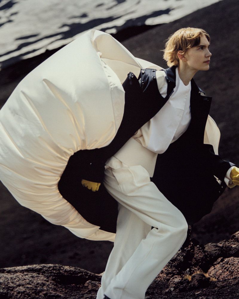 Fashion shot of a male model in a white outfit with an oversized white jacket against a dark, rocky landscape, posing and looking away.
