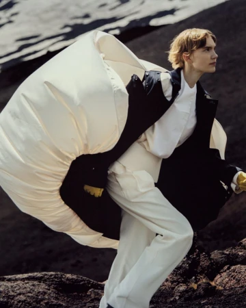 Fashion shot of a male model in a white outfit with an oversized white jacket against a dark, rocky landscape, posing and looking away.