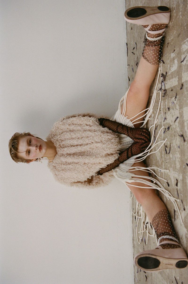 Fashion image of a young fair-skinned woman in a textured beige top, posing in a unique open-leg position with ballet flats and patterned socks.