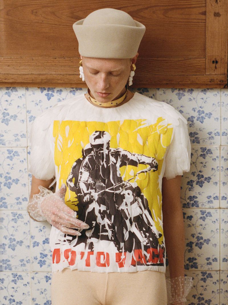 Fashion editorial shot of a fair-skinned person wearing a cream-colored hat and graphic tee with a motorcycle image, indoors.