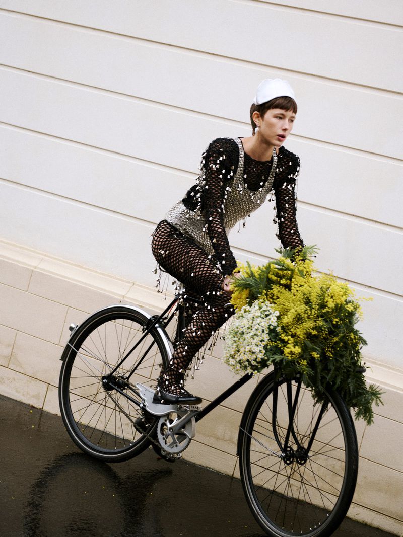 Fashionable woman in fishnet dress and cap rides a classic bicycle with a basket full of flowers against a textured white wall.