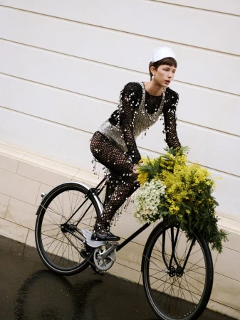 Fashionable woman in fishnet dress and cap rides a classic bicycle with a basket full of flowers against a textured white wall.