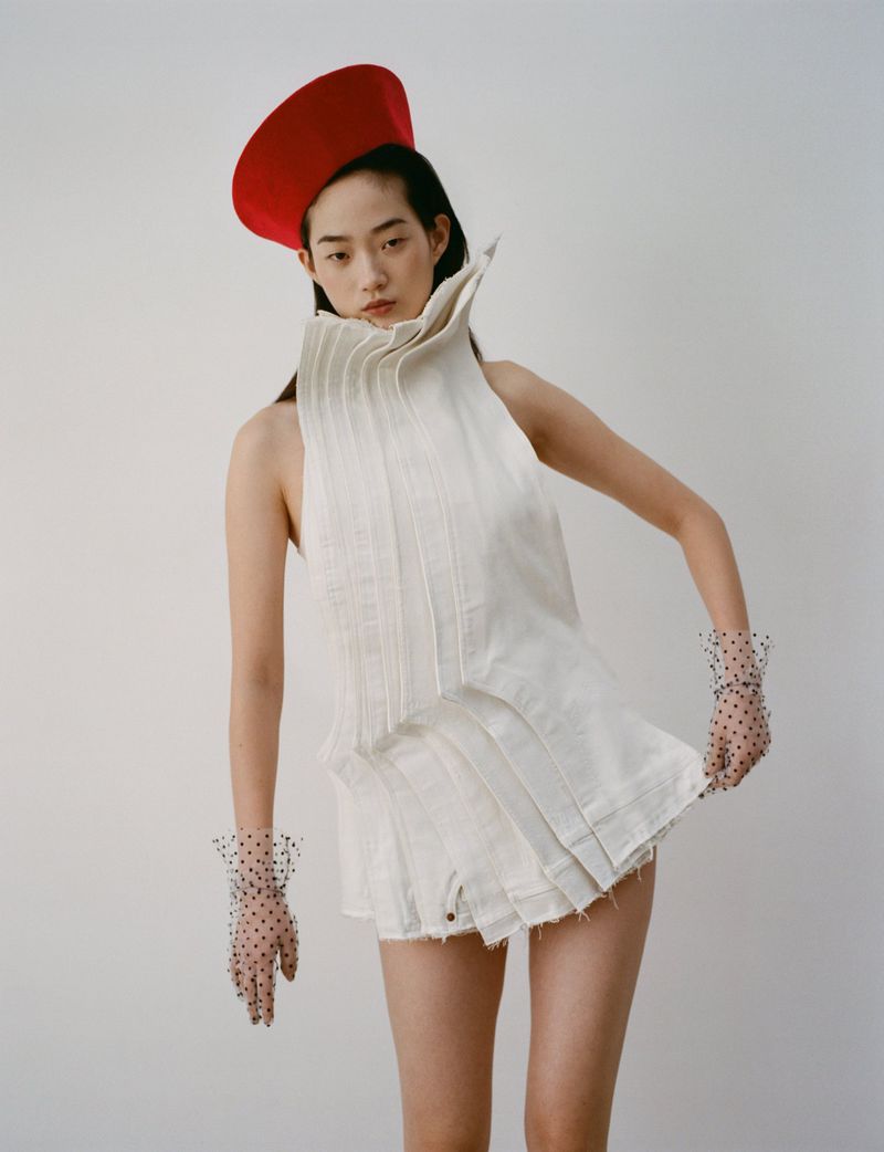 Editorial fashion shot of an Asian woman in a white pleated outfit with a red hat, posing in a studio against a plain white background.
