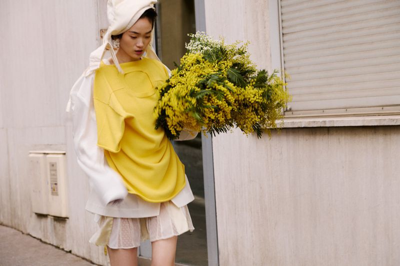 Asian woman with mimosa bouquet in off-white and yellow layers stands against a weathered building. Fashion-forward editorial shot.