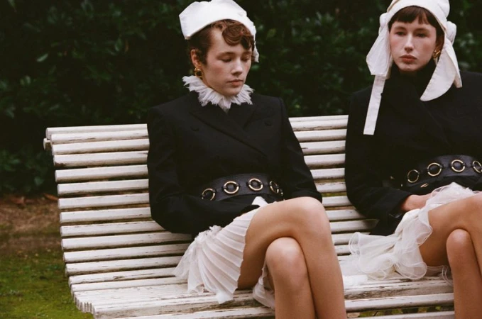 Two women in black blazers and white hats sit on a park bench, legs crossed. A lush green background creates a timeless fashion shot.