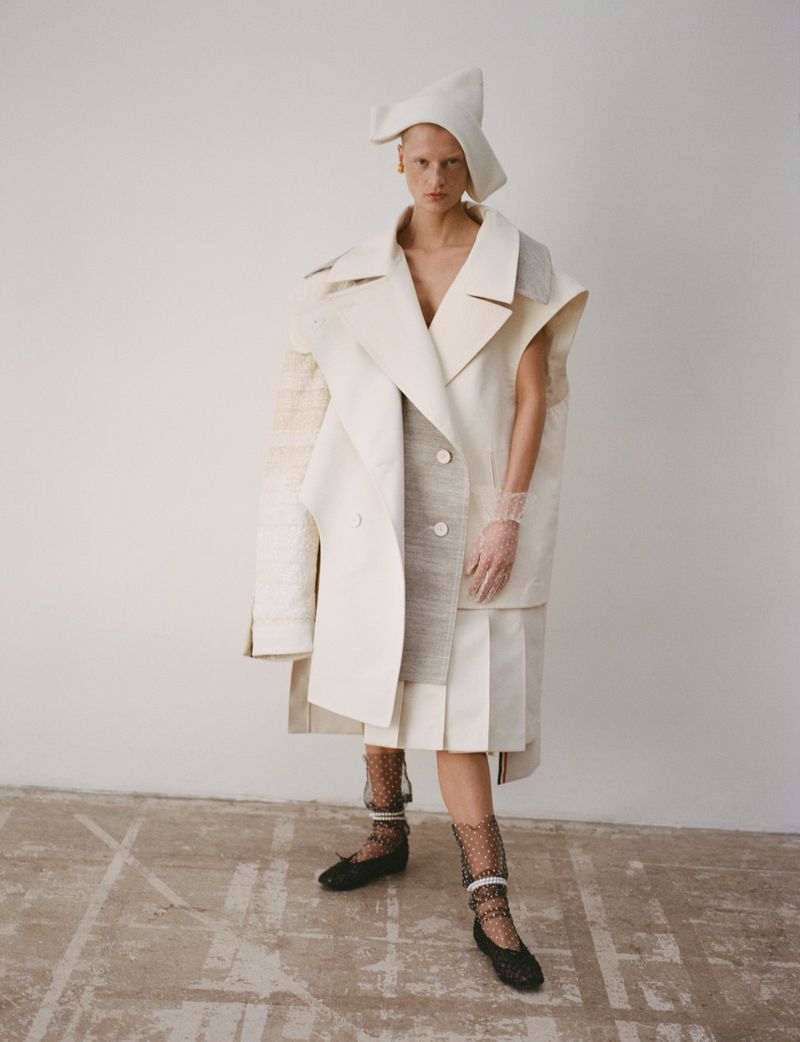 Fashion model poses in a white, sculptural coat with a matching pointed hat against a neutral backdrop in a studio fashion shot.