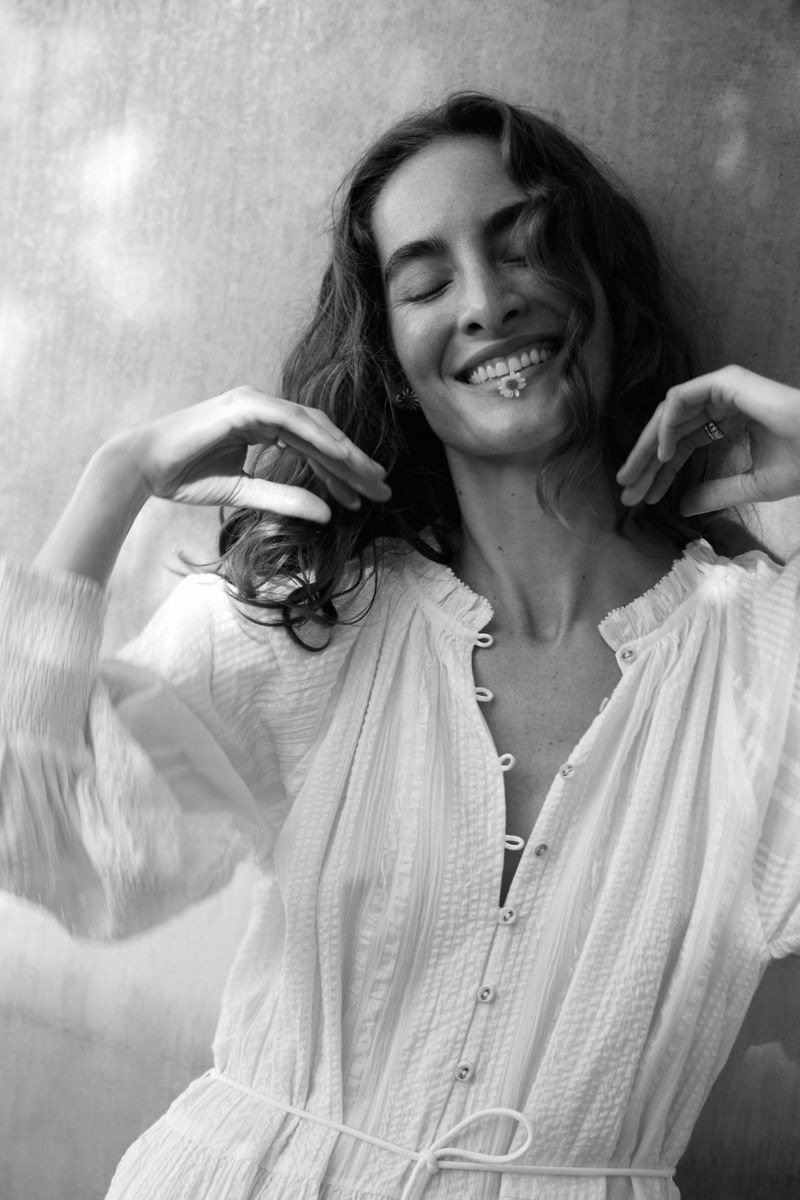 Woman with flower in her mouth smiles against textured wall in black and white, wearing a long-sleeved, buttoned, cotton dress.
