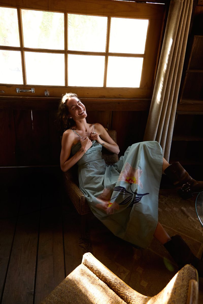 A woman in a long dress and boots sits smiling in an indoor, wood-paneled room with sunlight streaming through a gridded window.