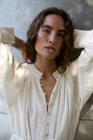 Portrait of a woman with dark hair, wearing a white dress, and holding a flower in her mouth, posing in front of a gray wall.