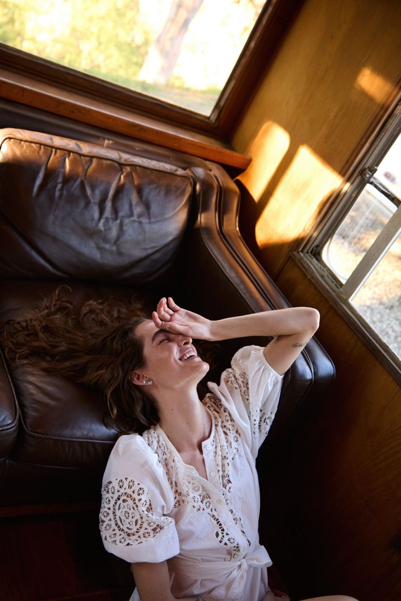 Woman in a white dress lounges on a leather couch near a window, smiling with a hand over her eyes, with natural light.