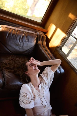 Woman in a white dress lounges on a leather couch near a window, smiling with a hand over her eyes, with natural light.