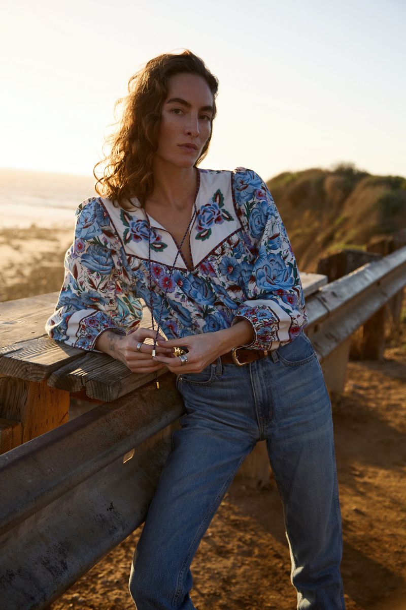 A young woman with long hair poses outdoors wearing a long-sleeved floral top, blue jeans, and a belt. The setting is sunny and natural.