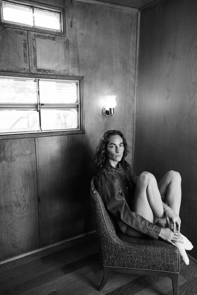 Monochrome portrait of a slender adult female with long wavy hair sitting on a patterned chair in a wood-paneled interior with windows.