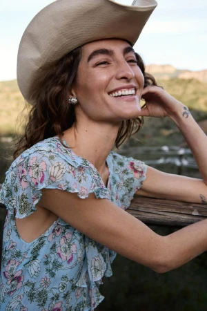 Smiling woman in a floral dress and cowboy hat stands near a wooden fence against a scenic mountain backdrop in an outdoor setting.