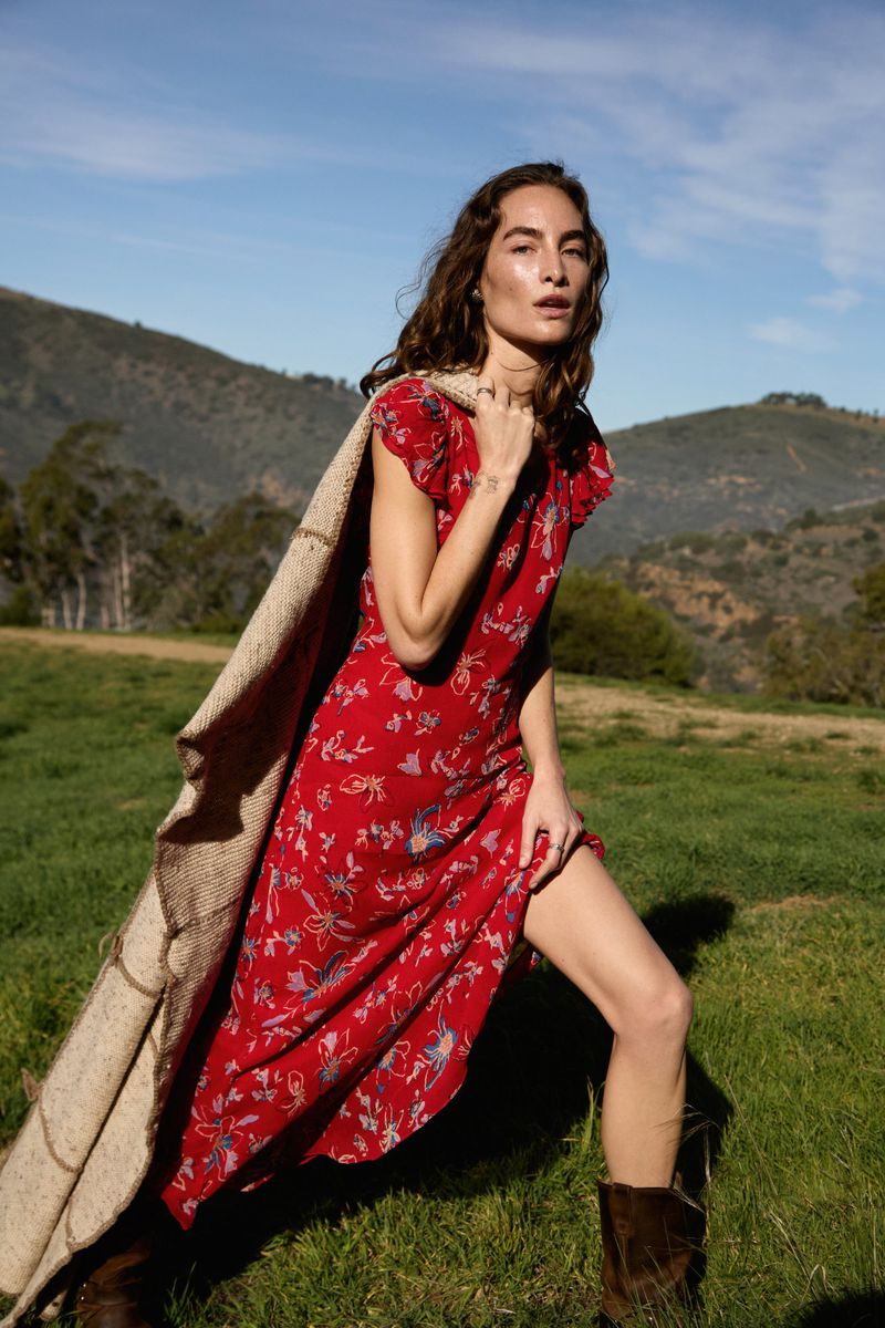 Model in a red floral dress holding a knit blanket outdoors in a grassy field with rolling hills and clear blue sky.