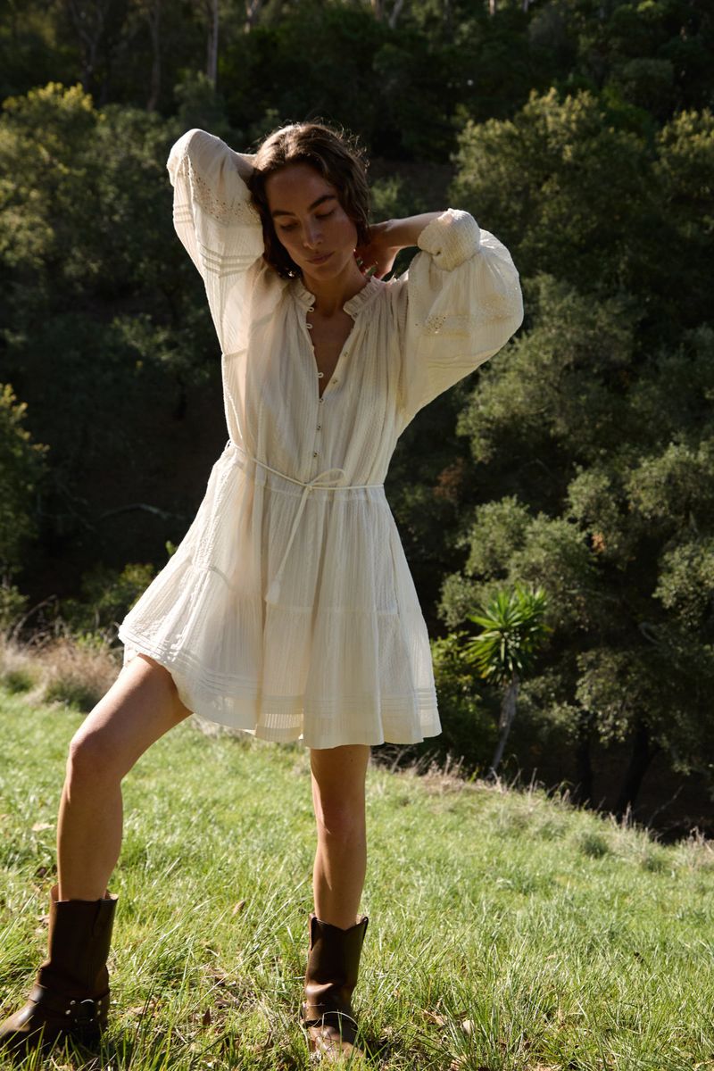 A woman in a short white dress and boots poses in a grassy field surrounded by trees. The image is shot in natural light.