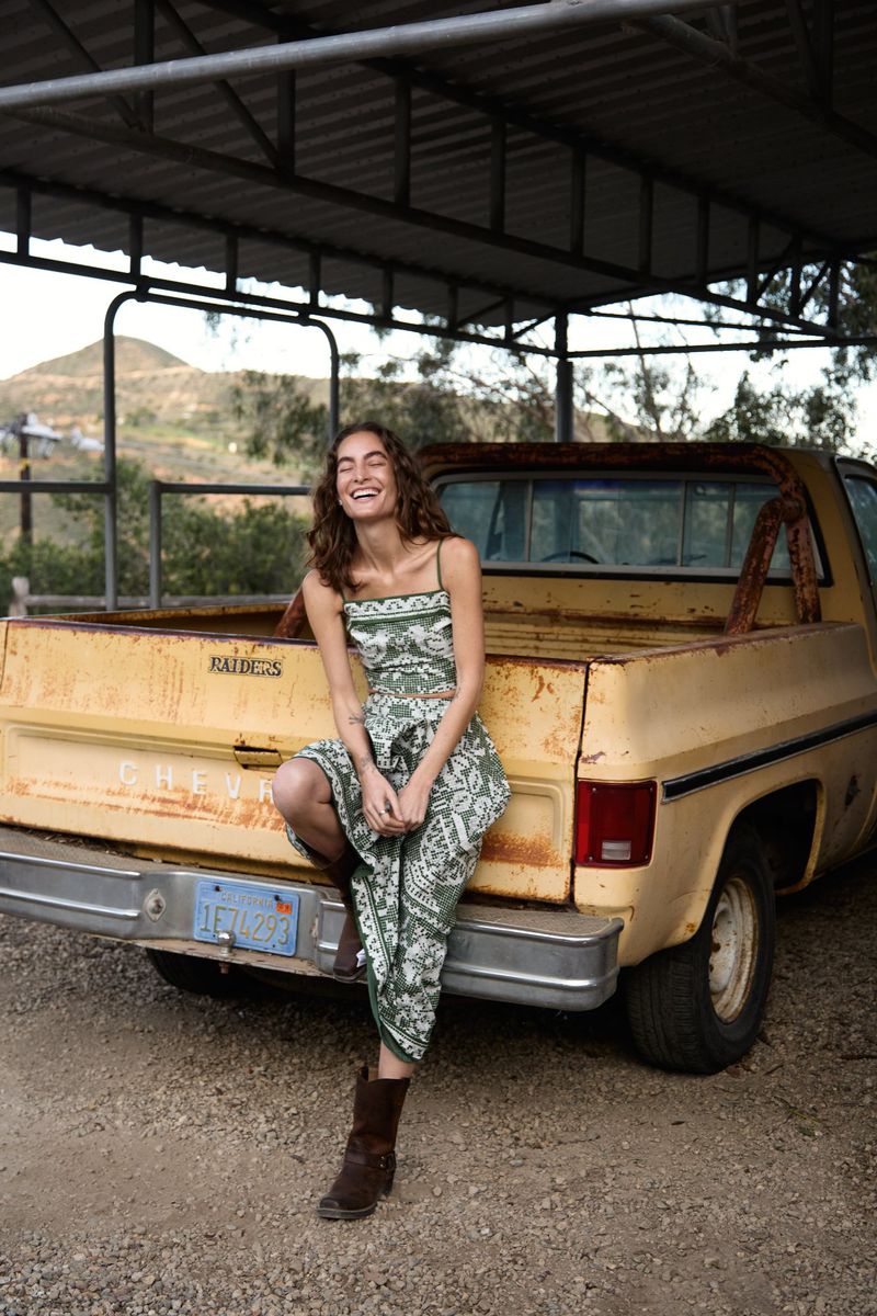 Woman in paisley dress poses on vintage yellow pickup truck under covered structure with mountains visible in background on sunny day.