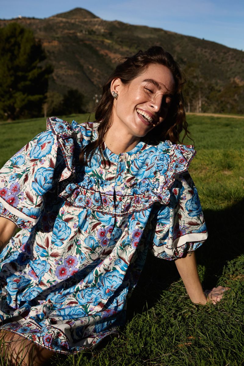 Woman wearing a blue floral dress laughing while seated on a grassy hillside under a clear sky.
