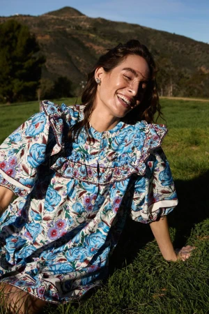 Woman wearing a blue floral dress laughing while seated on a grassy hillside under a clear sky.