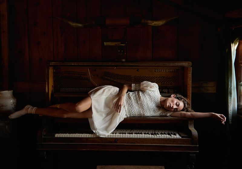 Woman in white dress poses on an antique piano, with wooden walls and steer horns creating a rustic yet elegant, musical atmosphere.