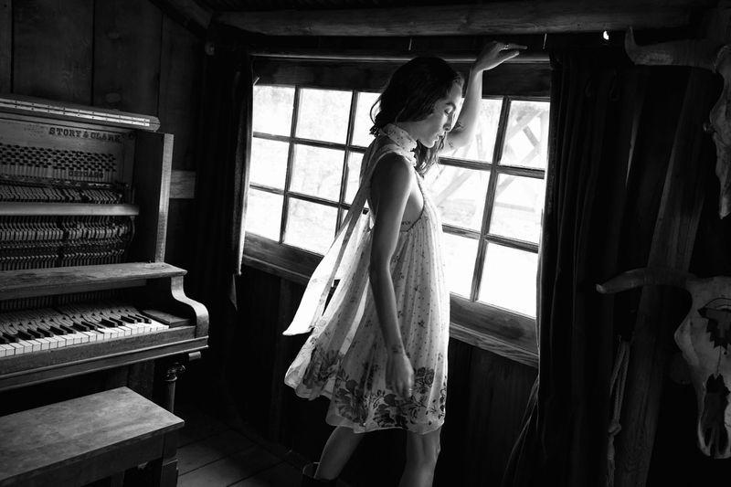 Monochrome shot of a slender woman in a patterned dress standing by a window in a rustic room with a vintage piano.