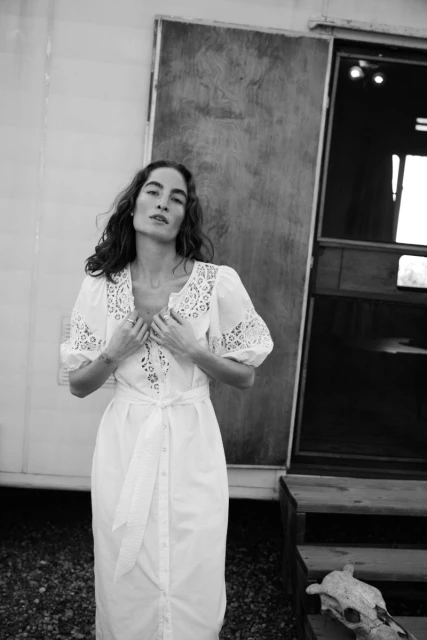 Monochrome portrait of an adult woman in white eyelet dress standing outside a trailer with a steer skull nearby, in medium shot.