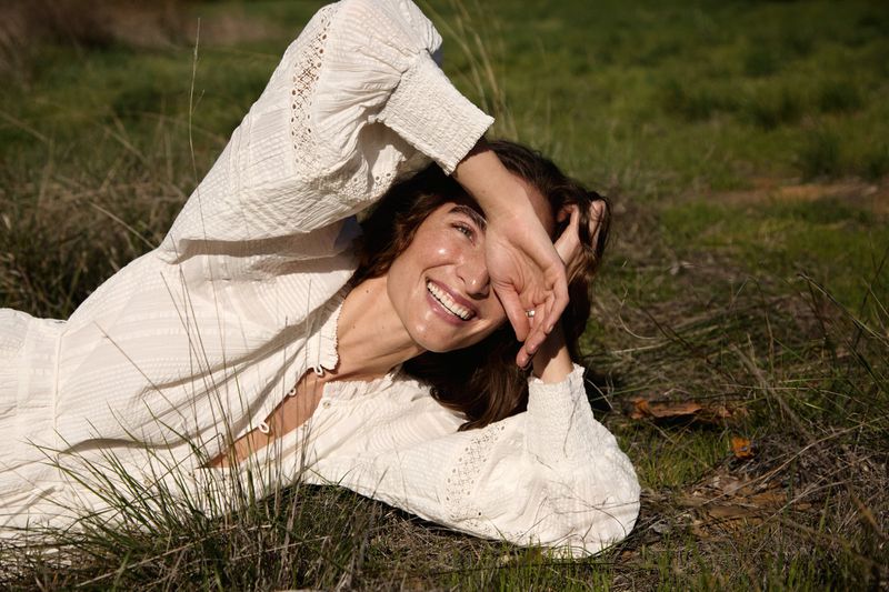 Woman in a white dress lies in a grassy field, smiling and shielding her eyes from the sun, creating a joyful, relaxed outdoor portrait.