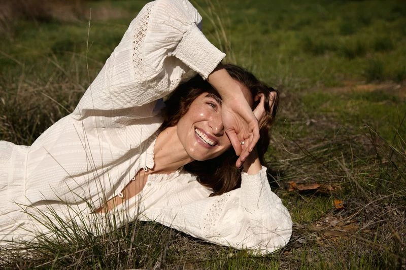 Woman in a white dress lies in a grassy field, smiling and shielding her eyes from the sun, creating a joyful, relaxed outdoor portrait.