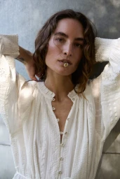 Model Heather Kemesky holding a small flower in her mouth wearing a white textured blouse against a textured stone wall
