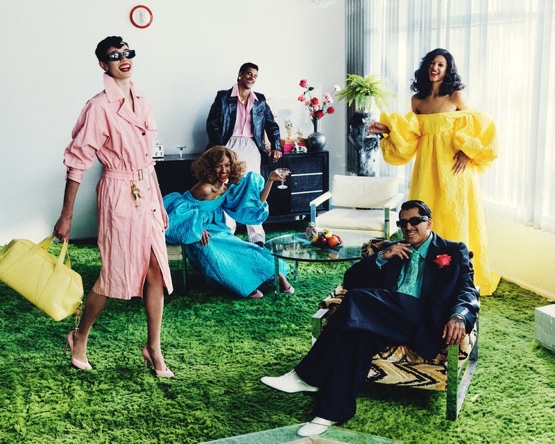 Five people in vibrant vintage attire pose indoors with drinks and stylish furniture, set against a light wall and green shag carpet backdrop.