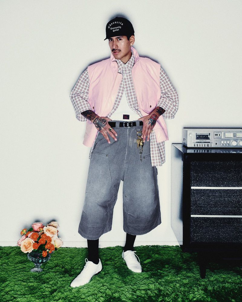 A man in a pink vest and baggy jeans poses in a photo studio with a retro aesthetic featuring green carpet and a vintage stereo.