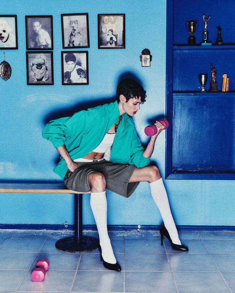 A woman in a green jacket sits on a bench lifting a pink dumbbell in a blue room with trophies and framed photos on the wall.