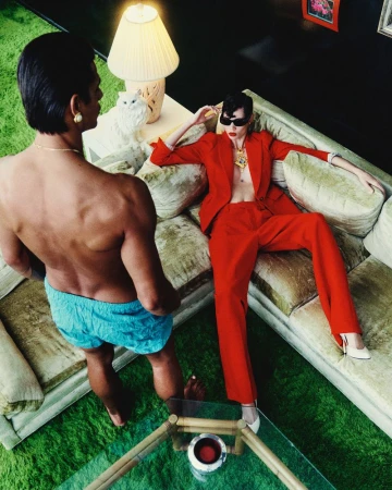 High-angle shot of woman in red suit reclining on a sofa, near a shirtless man in swim trunks, indoor living room setting with green carpet.