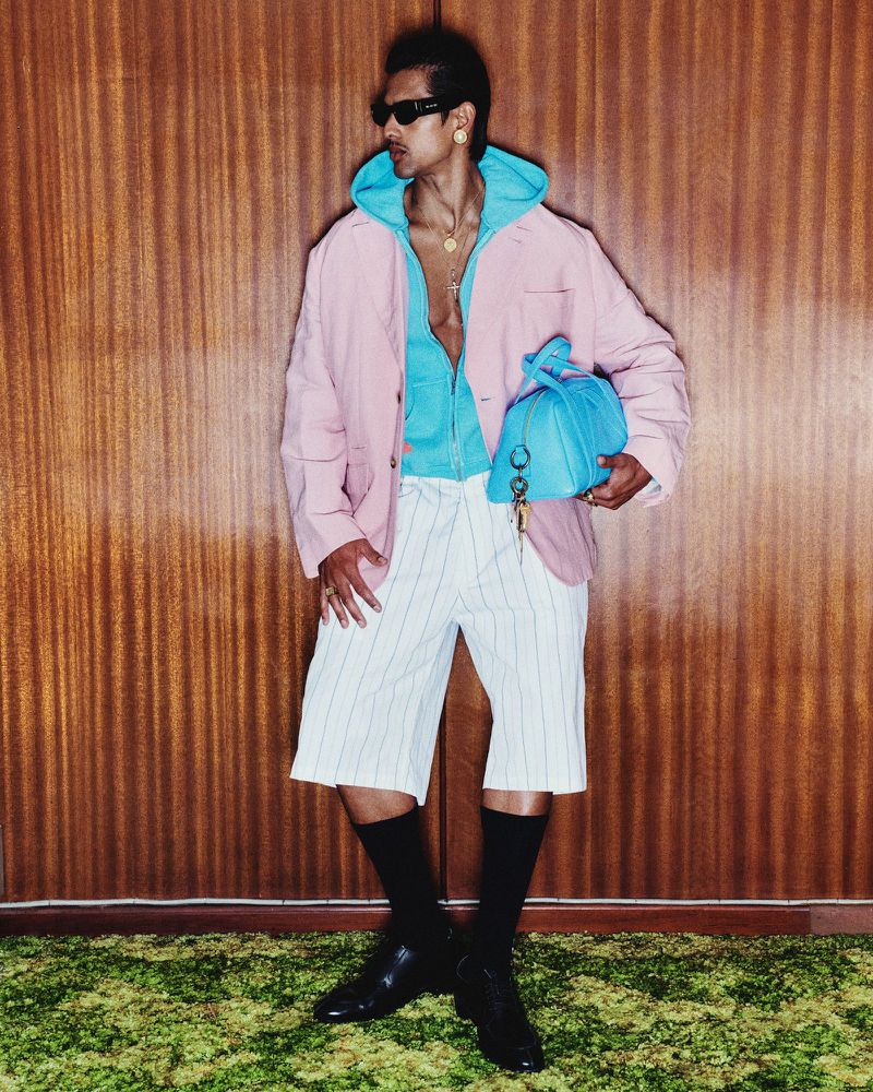 Adult male poses in a pink blazer, light blue hoodie, and pinstripe shorts, holding a blue bag in an indoor studio setting.