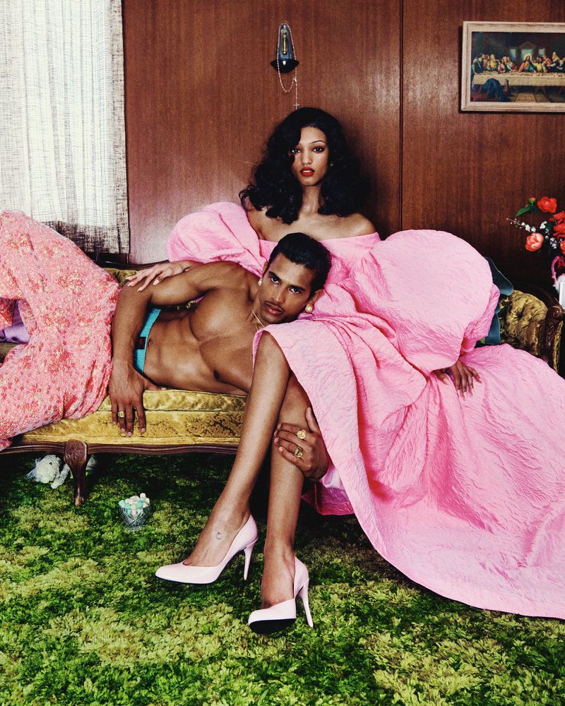 Two models draped in pink fabric pose in an interior setting. One is a shirtless man, the other an African-American woman.