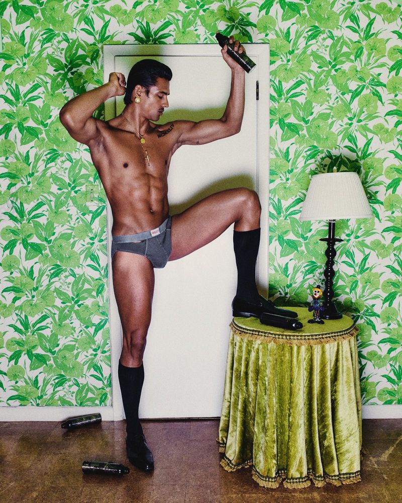A man in underwear strikes a pose next to a lamp, with a leaf-patterned wallpaper in the background, and several objects on the floor.