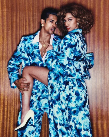 Glamorous couple in matching blue floral suits strike a pose against a brown wooden backdrop. Woman wears white heels.