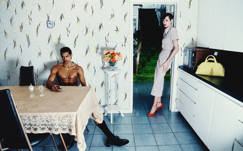 A man sits at a table in a kitchen across from a woman leaning against a counter. The kitchen features patterned wallpaper.