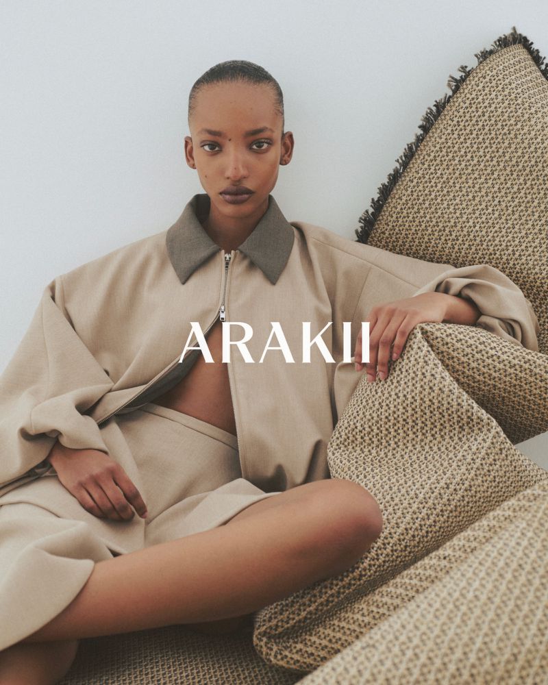 A Black female fashion model poses indoors in a beige jacket and shorts, seated on woven pillows, with the brand ARAKII in the foreground.
